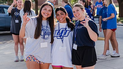 Three new LCU students holding up the Chap hand sign as belongings are moved into a dorm behind them