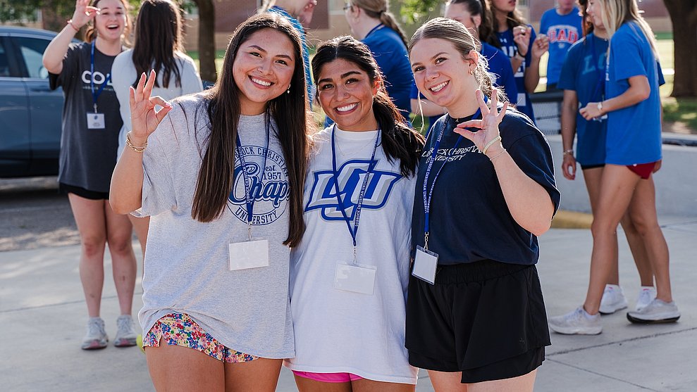Three new LCU students holding up the Chap hand sign as belongings are moved into a dorm behind them