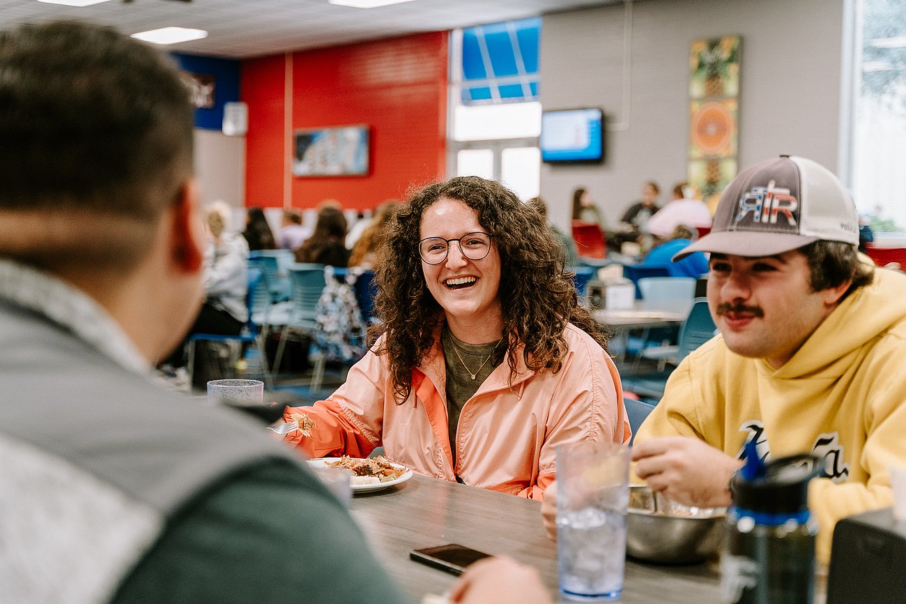 A picture of three students sitting in the cafe eating their food as two of the students look and laugh at the student sitting across from them in enjoyment of the conversation