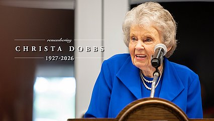 Photograph of Christa Dobbs behind a lectern and the words, "Remember Christa Dobbs: 1927-2026"