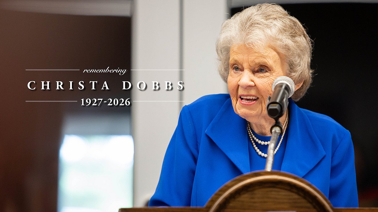 Photograph of Christa Dobbs behind a lectern and the words, "Remember Christa Dobbs: 1927-2026"