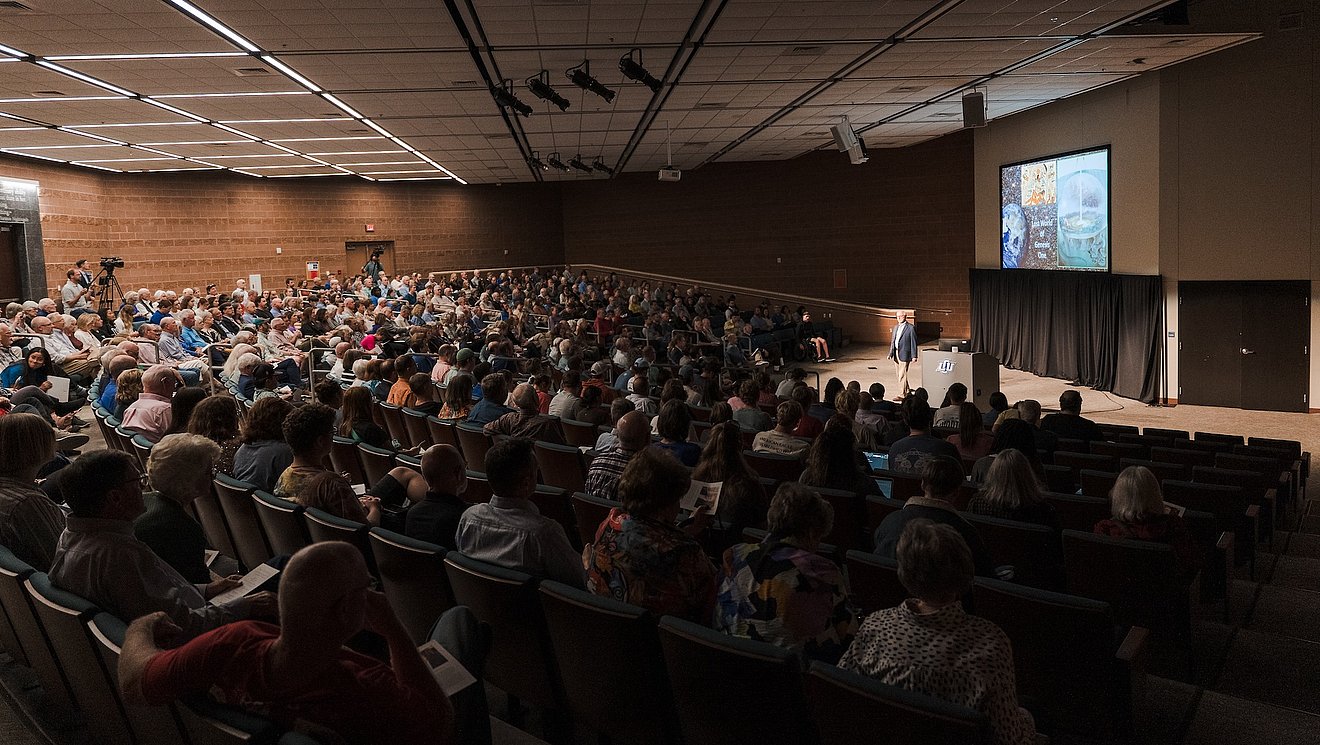 Photograph of crowded room of attendees listening as Dr. Walton gives lecture on stage