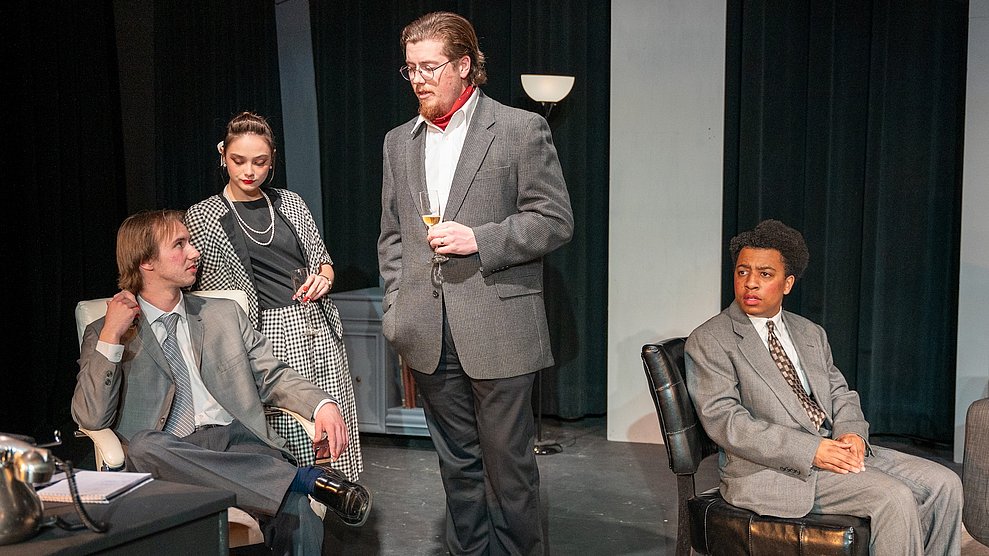 The cast of Murder by the Book in a scene in mid-century dress with some sitting on a couch and one standing beside in coversation