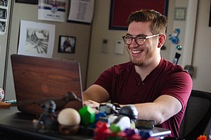 A smiling man in glasses and a red shirt is focused on his laptop in an office setting