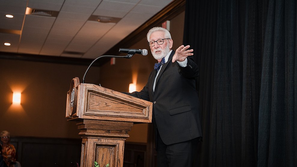 Photograph of Doc Williams in a suit speaking behind a lectern on stage at the dinner