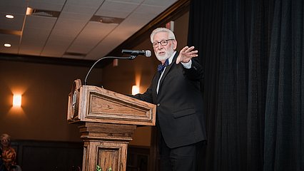 Photograph of Doc Williams in a suit speaking behind a lectern on stage at the dinner
