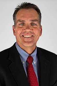 Professional portrait of Toby Rogers smiling towards the camera in a black suit with a blue shirt and red tie