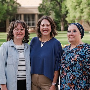 Photograph of three women, all employees of the student business office, standing outside and smiling at the camera