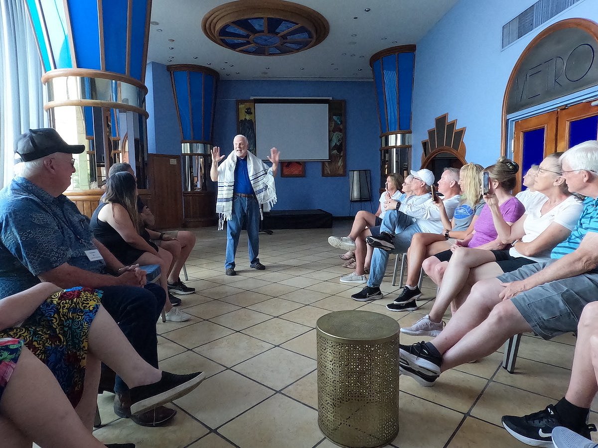 A group of people sitting along the left and right wall looking at a man in a prayer shawl in a blue room with gold accents