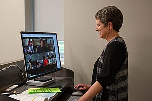 A woman standing at a podium with her notes in front of her on a video call, wearing a black and gray shawl and a smile as she talks to her colleagues