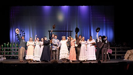 Stage photograph of the cast of OKLAHOMA! singing at the end of the show in western costumes and dramatic stage lighting