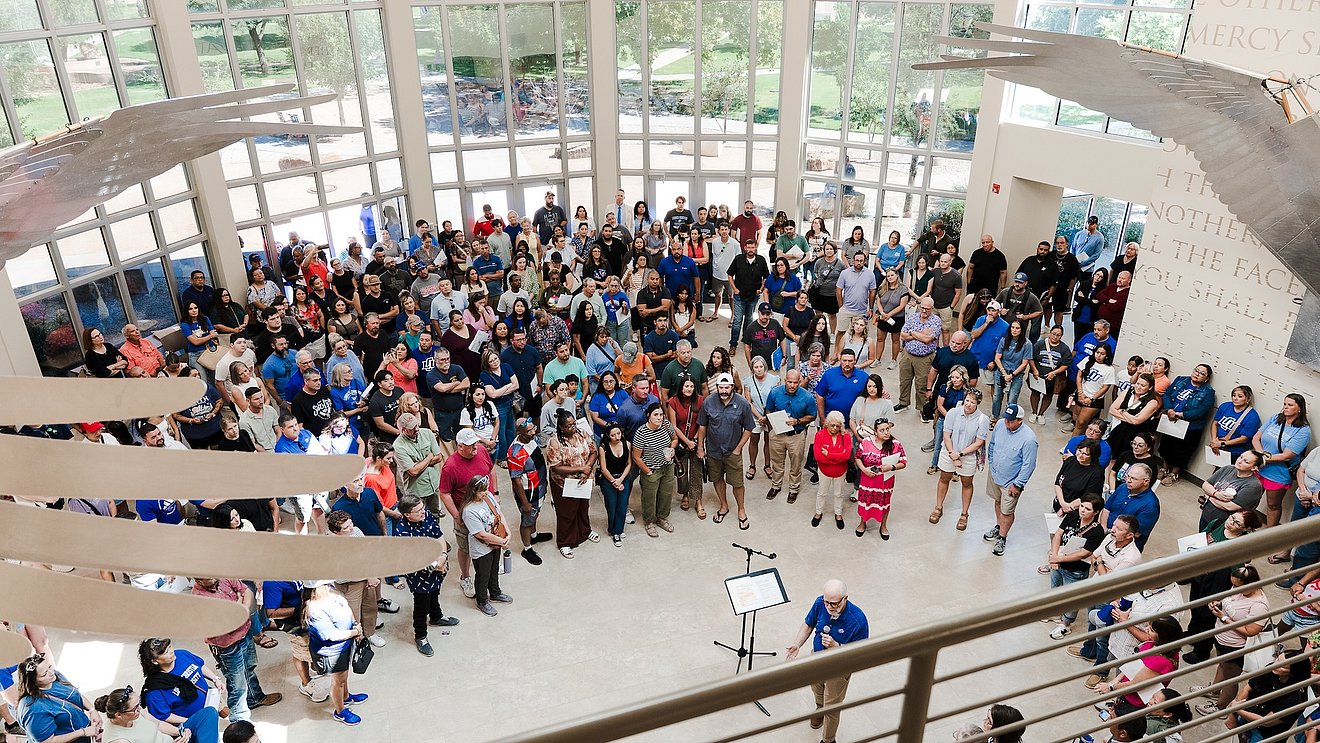 overhead photograph of LCU freshmen students being blessed by faculty beneath an indoor sculpture of the cherubim's wings over a large foyer area with a wall of windows in the background