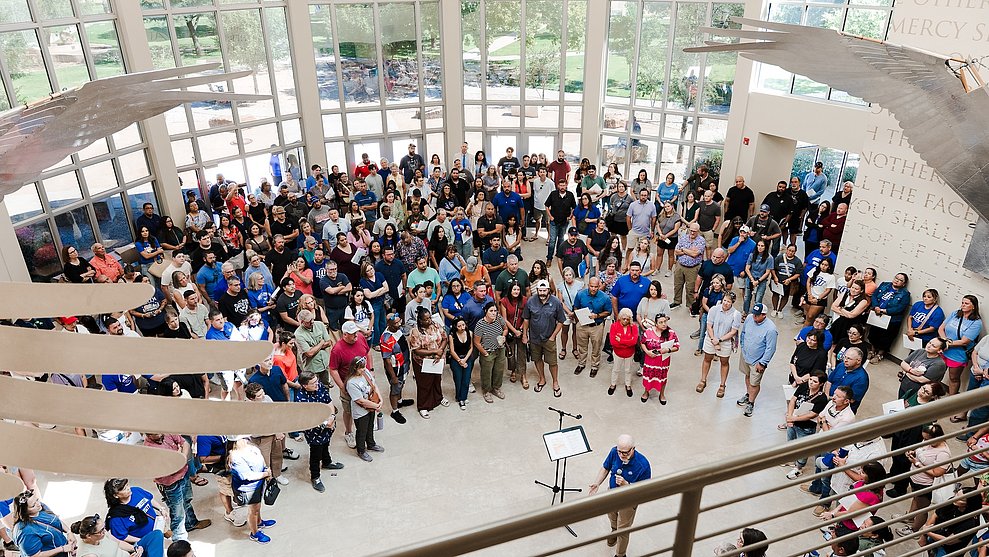 overhead photograph of LCU freshmen students being blessed by faculty beneath an indoor sculpture of the cherubim's wings over a large foyer area with a wall of windows in the background