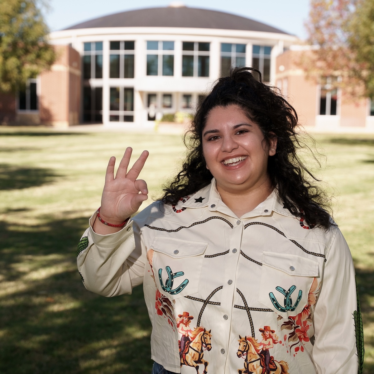 Photograph of Liliana Jiminez-Cuello holding up the chap hand sign in front of the Welcome Center at LCU