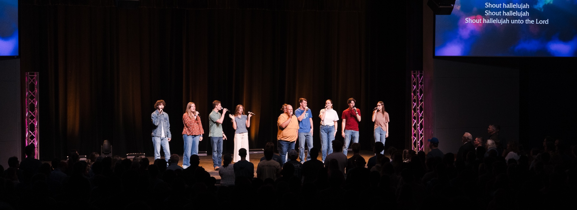 Photograph of the inside of the McDonald Moody Auditorium from behind audience looking at stage, dark lights, worship team on stage, lyrics for a worship song on screens with dramatic lighting