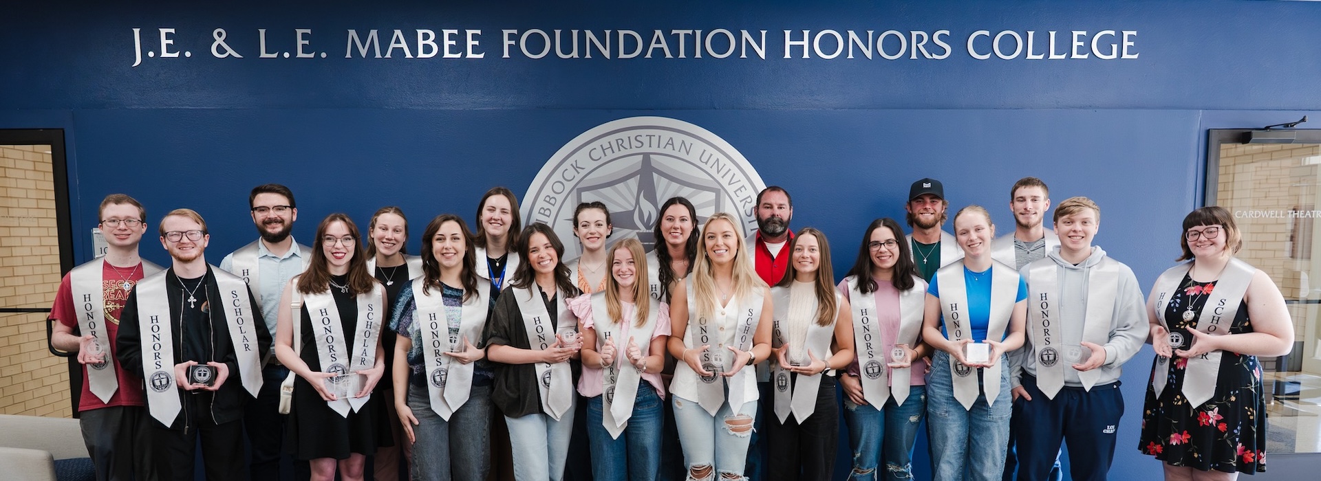 Photograph of students in the common area of the Mabee Foundation Honors College all wearing honors sashes and holding crystal paperweights inscribed with the honors college logo