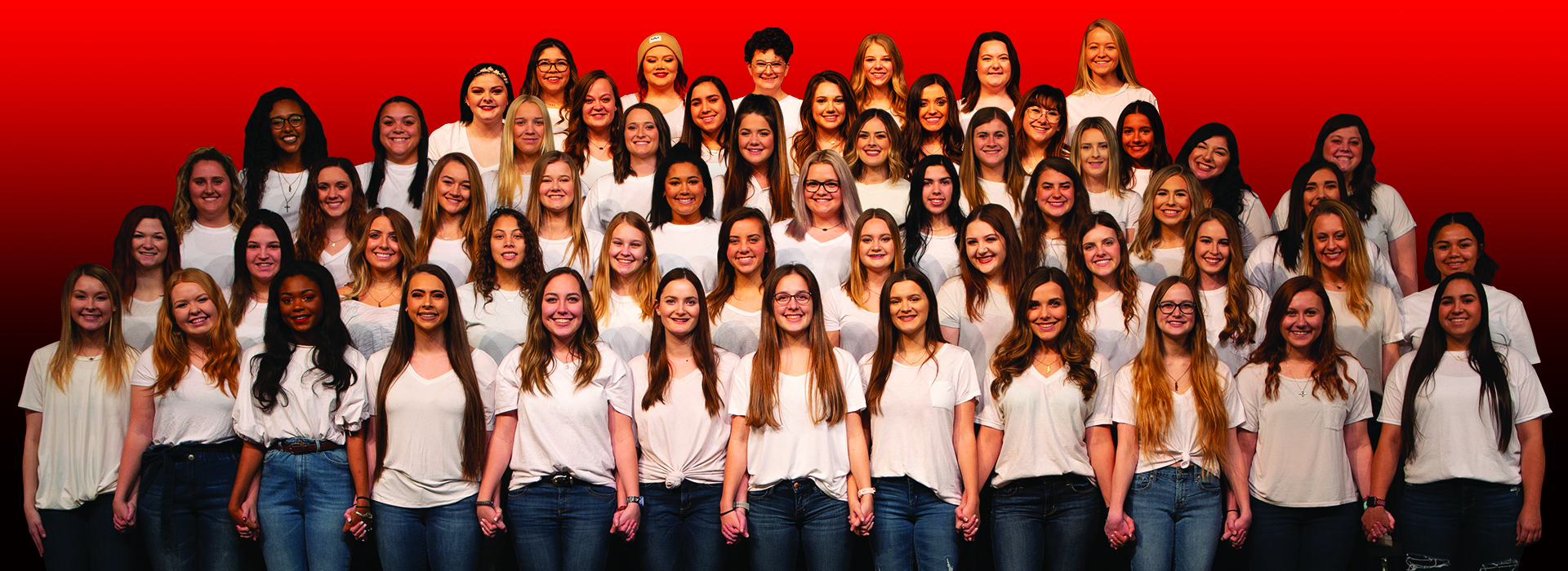 A group of girls in Kappa Phi Kappa standing on risers, in white shirts smiling towards the camera, with the front row holding each others hands 