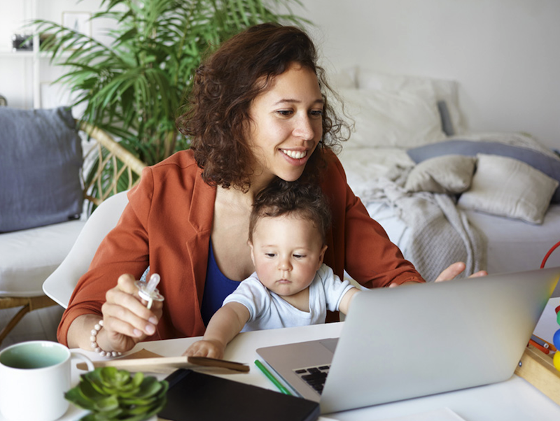 Smiling woman sitting at her desk with laptop working while holding a toddler in her lap