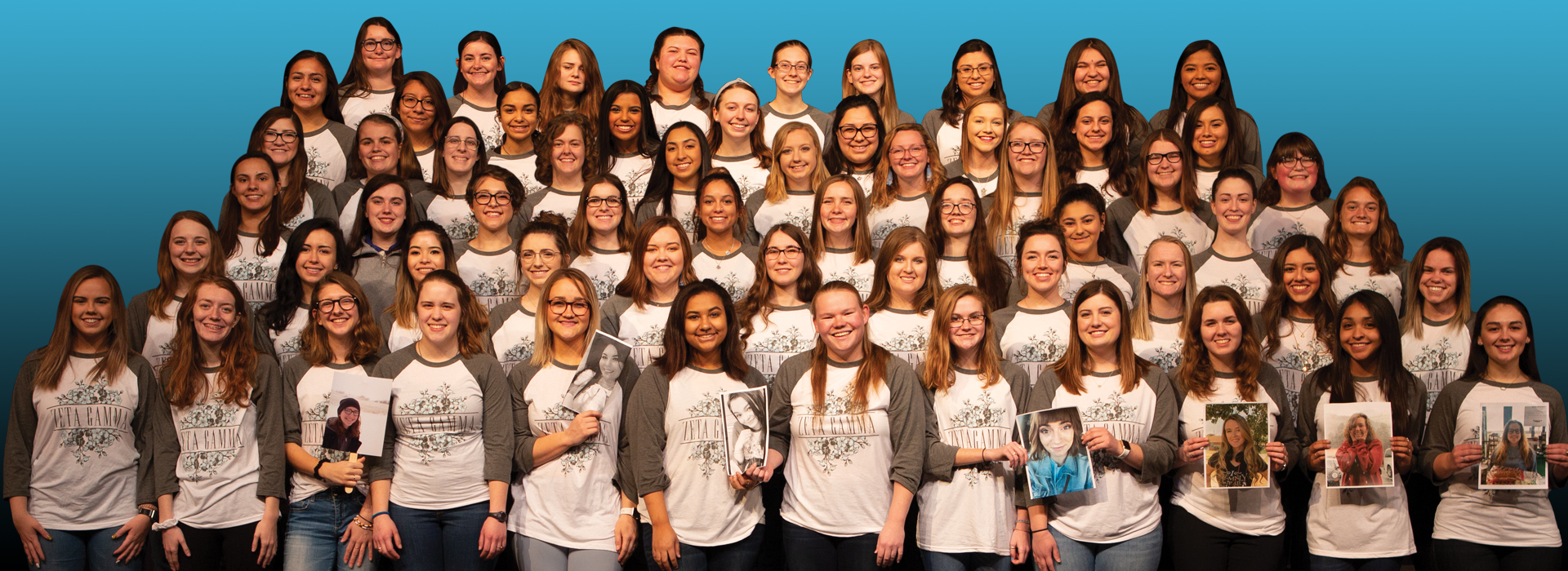 A group of girls in Zeta Gamma wearing a Zeta Gamma shirt standing on risers and smiling towards the camera, in the front row, you can see seven girls holding a picture of the girls who could not make it in the picture